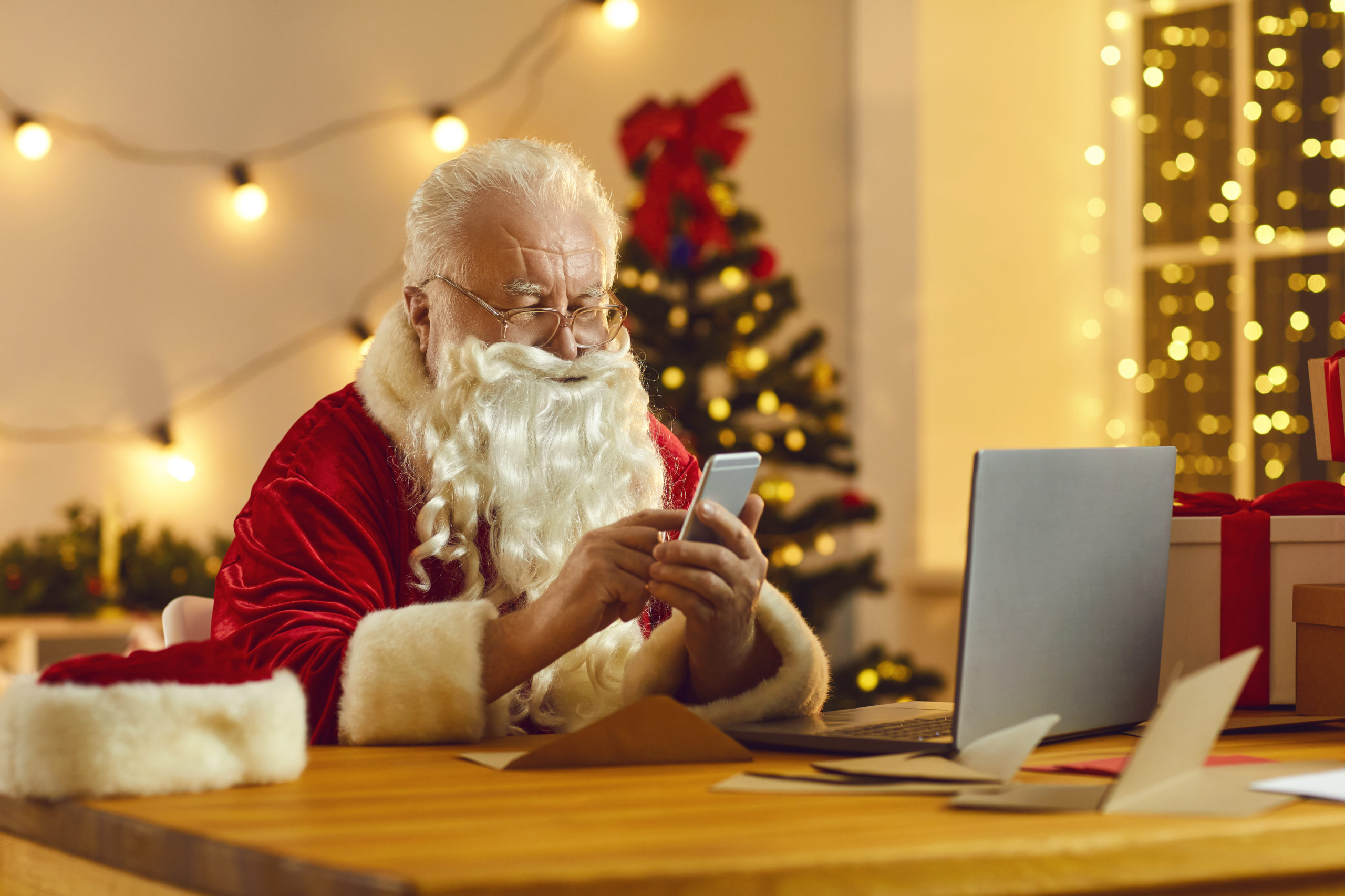 Modern Santa Claus looking at mobile screen, checking his email box and reading letters from children. Father Christmas sitting at desk in his workshop using phone app to order presents delivery