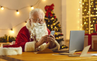 Modern Santa Claus looking at mobile screen, checking his email box and reading letters from children. Father Christmas sitting at desk in his workshop using phone app to order presents delivery